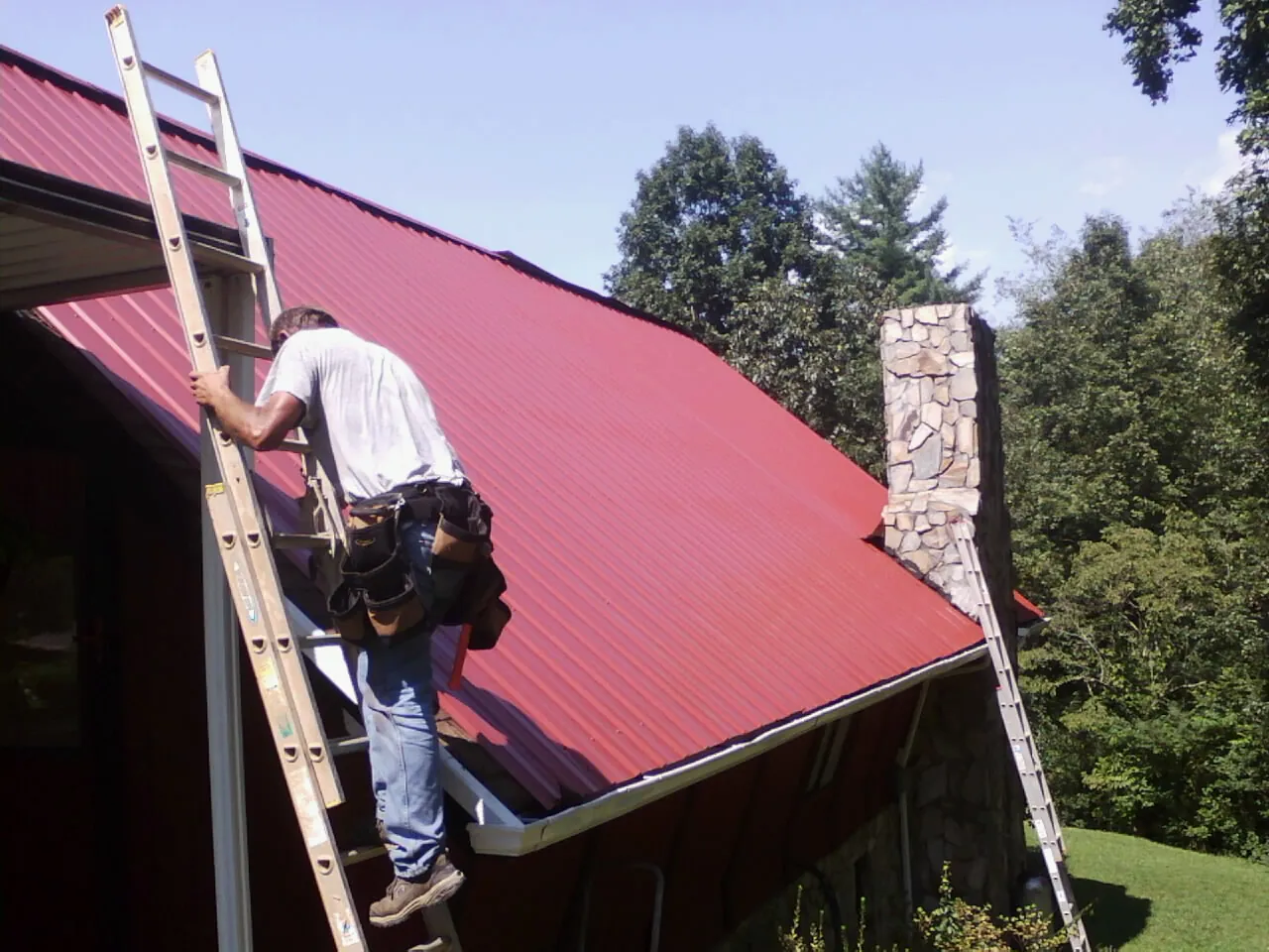 Roofer on a ladder beside a red metal roof and stone chimney — Soffit & Fascia Repair by Roofing Brothers in Port Charlotte